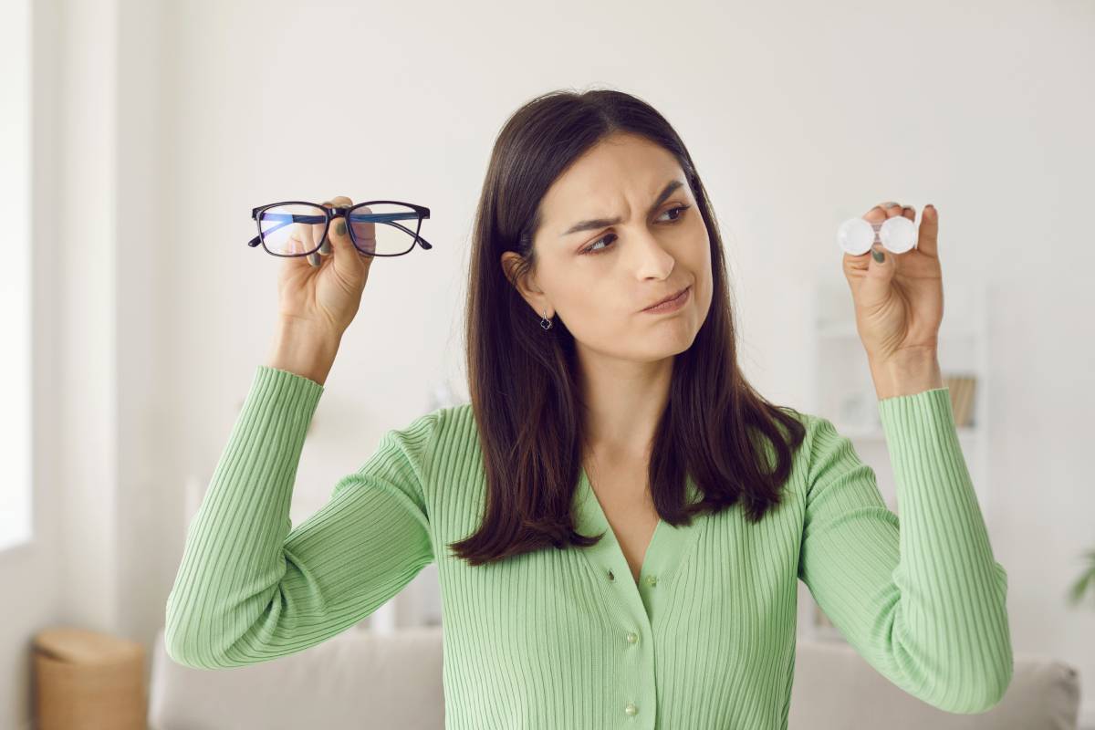 Portrait of pensive woman hesitating choosing between glasses and contact lenses. Young woman with questionable expression looks at contact lenses she holds in one hand and has glasses in other hand.